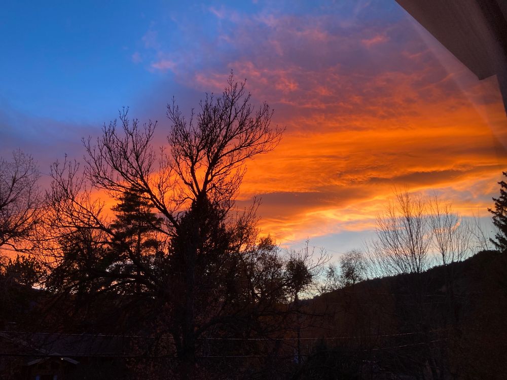 crazy orange sunset stretching across the dark backdrop of foothills to the west...a corner of blue sky is to upper left..bare trees are outlined by the sunset
