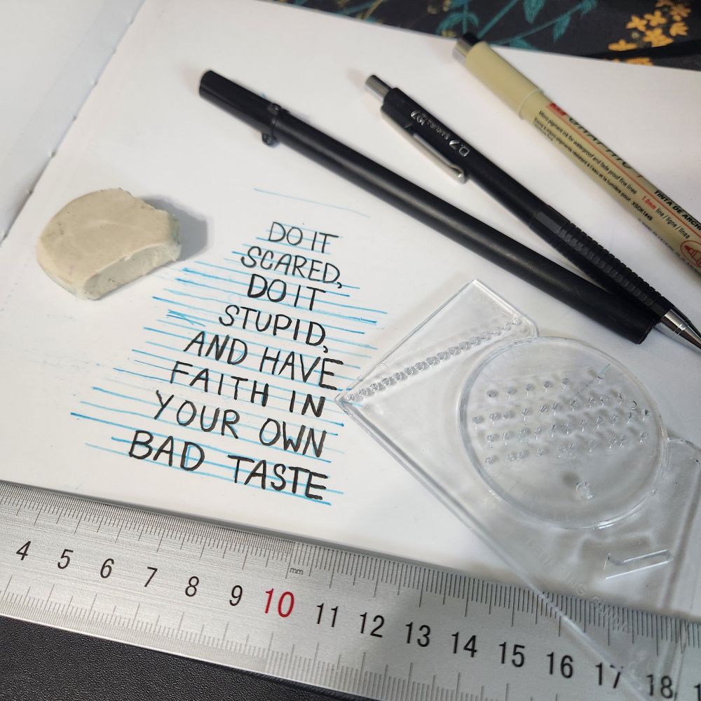 A sketchbook lying open on a desk, covered in drawing supplies. Black lettering drawn over blue guidelines reads, "Do it scared, do it stupid, and have faith in your own bad taste."