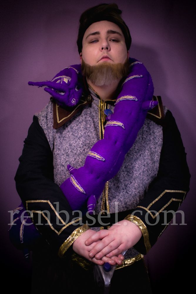 A photo of Ren Basel, a white nonbinary person, dressed in cosplay as Viren from The Dragon Prince. They have brown hair and a beard, wear courtly wizard attire, and have a plushie of a large purple worm draped across their shoulders. Their hands are rested on a staff.