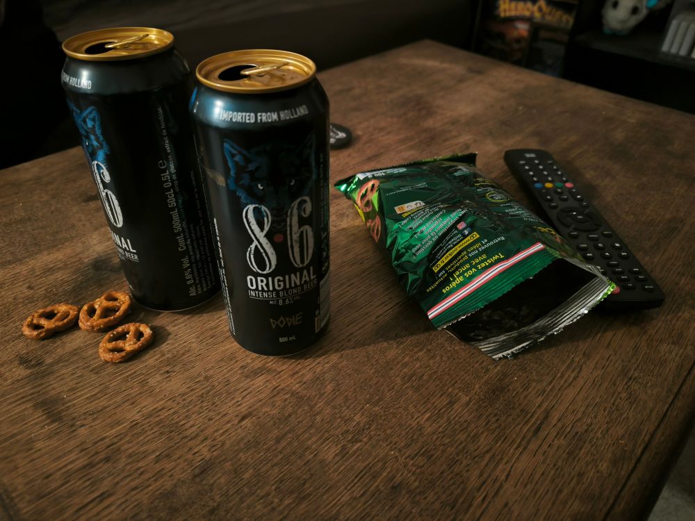 Une photo de deux bières de marque 8.6 avec un sachet de bretzel, à côté d'une télécommande sur une table en bois 