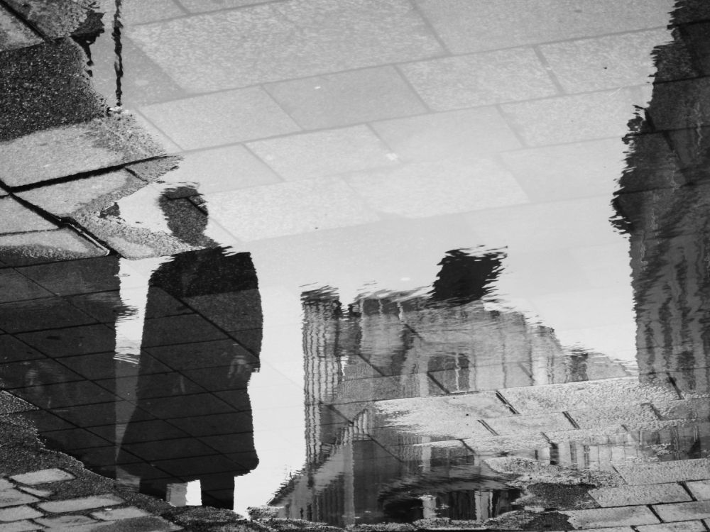 person in a black coat mirrored in a puddle near Stephansdom, with famous Haas house in the background (also upside down), black and white photo