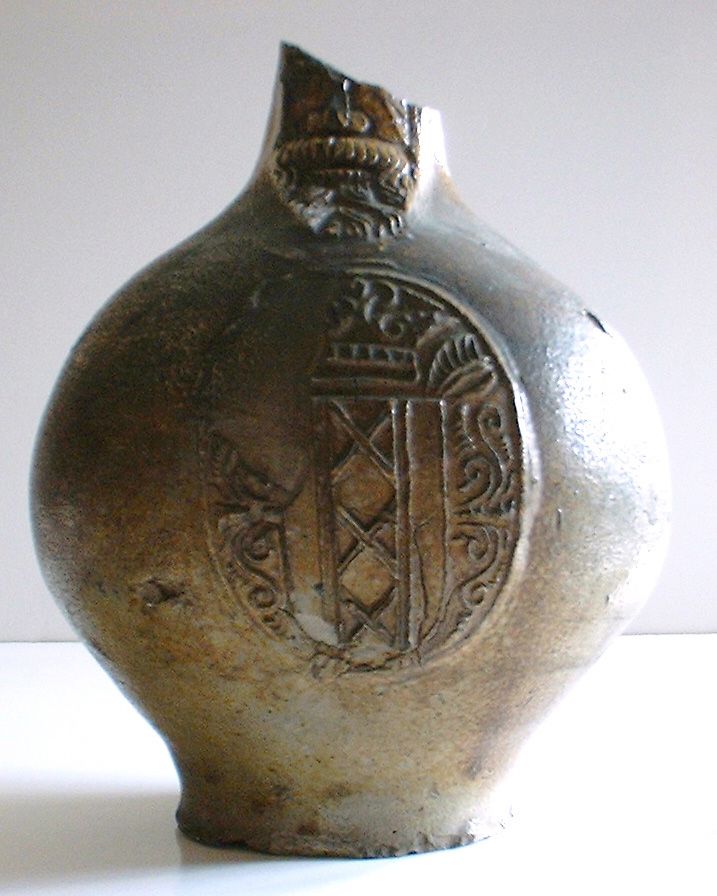 Brown glazed bellarmine jar with the handle and part of the neck missing. There is a well defined face of a bearded man decorating the neck. On the body there are the remains of an applied oval badge depicting a stylised coat of arms. There is a triangular ridge where the handle joined the body of the jar.