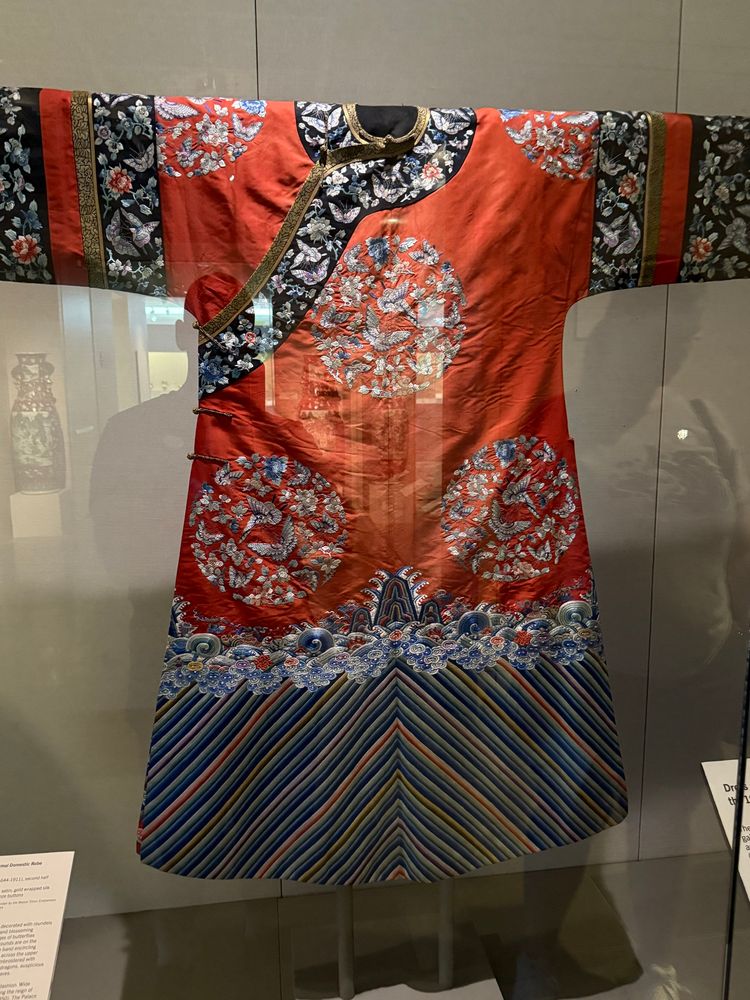 A woman’s silk robe from the Chinese Qing dynasty. The robe is bright red with embroidered flowers and other patterns and a striped blue bottom.