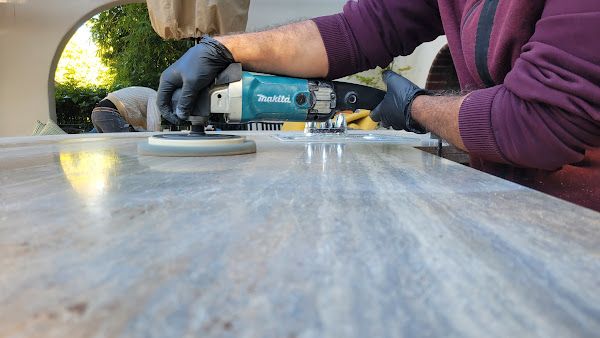 A professional technician polishing a natural stone surface using a Makita polisher. The reflection on the stone shows its smooth, glossy finish under outdoor lighting.