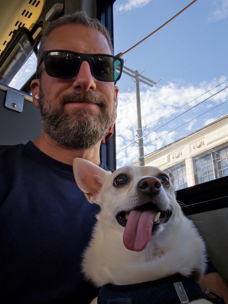 Matt sitting on the bus with his Chihuahua-Corgi mix Toby in his lap. Out the window it’s a nice partly cloudy day with blue skies.