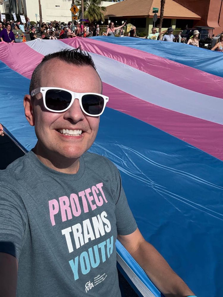 Scott wearing a tshirt that reads protect trans kids while holding onto a gigantic trans pride flag in a parade. 
