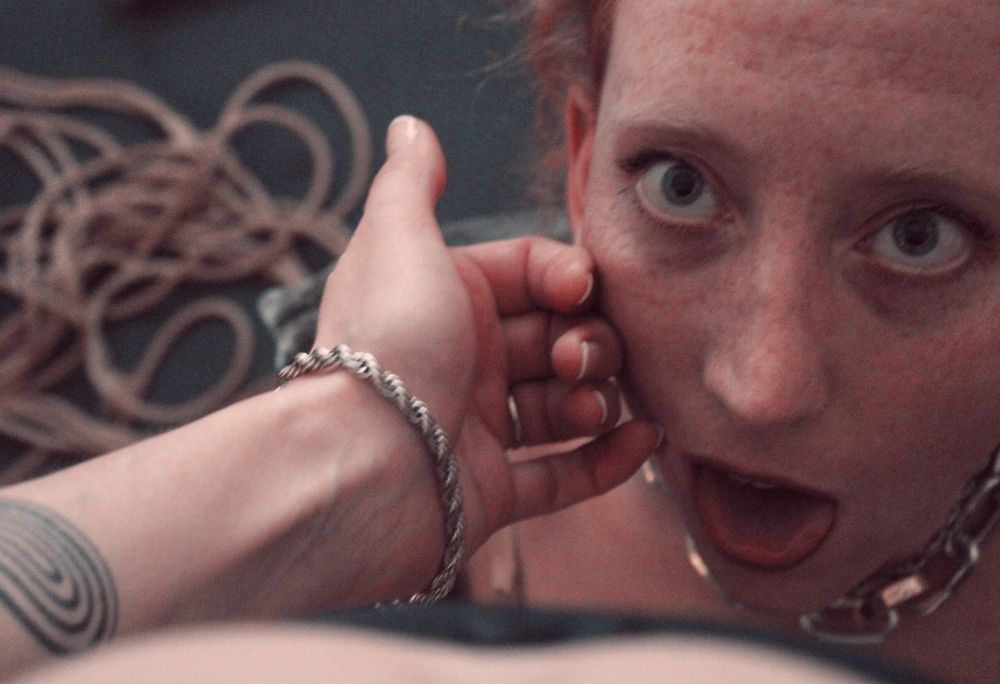 Close up of Rhiannon who is kneeling facing the camera with her tongue out and eyes wide, ready for a gift from her Madam, who gently strokes her cheek.