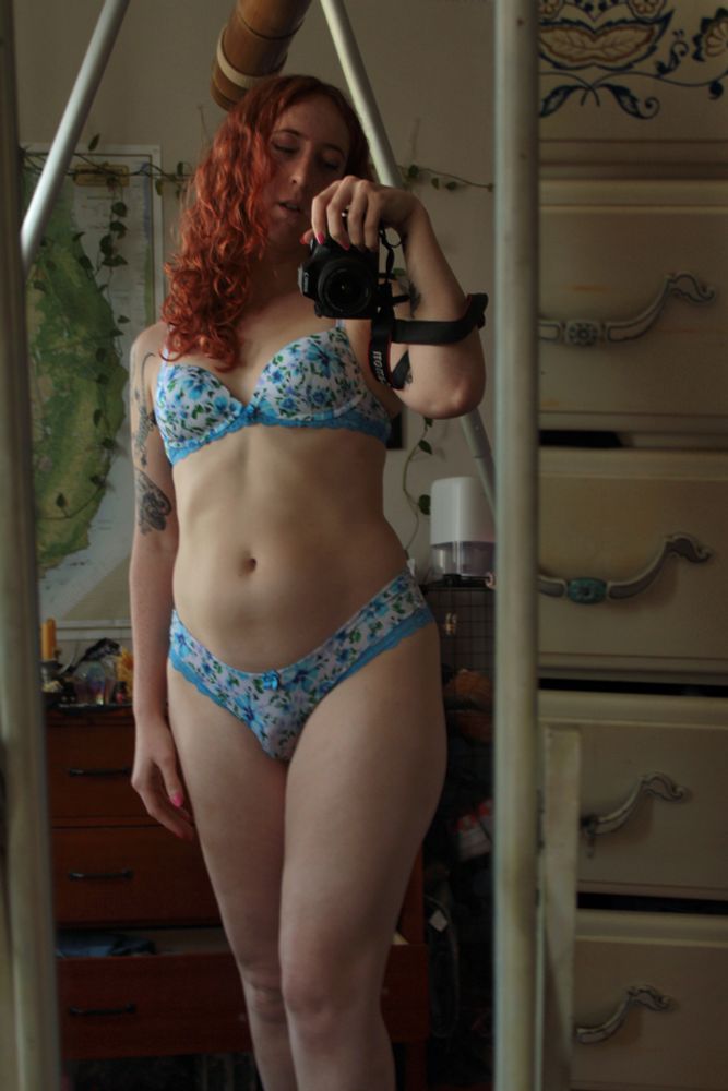 Rhiannon stands wearing blue flowery lingerie, taking a mirror selfie with a camera.