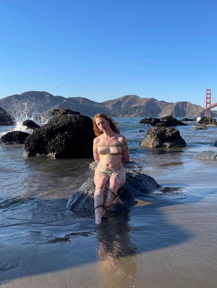 Rhiannon sits calmly on a rock by the sea, tied in rope with only her panties on. A wave crashes on a nearby rock.