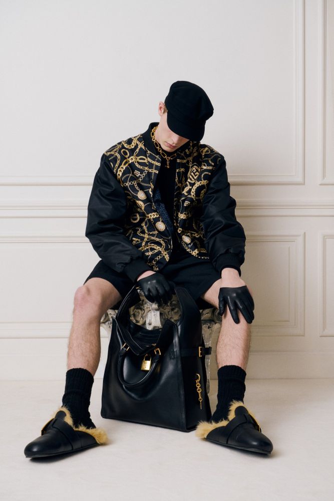 White male model wearing a black outfit with fold accent, a hat, gold jewelry, and furry and leathery slippers