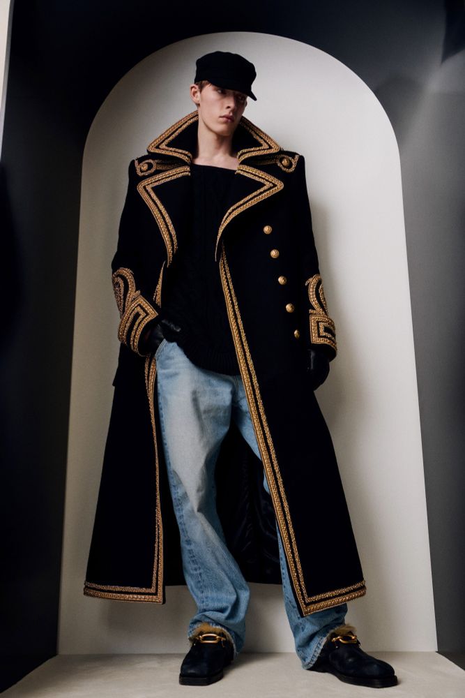 White male model wearing a black and gold coat with a hat and blue jeans 