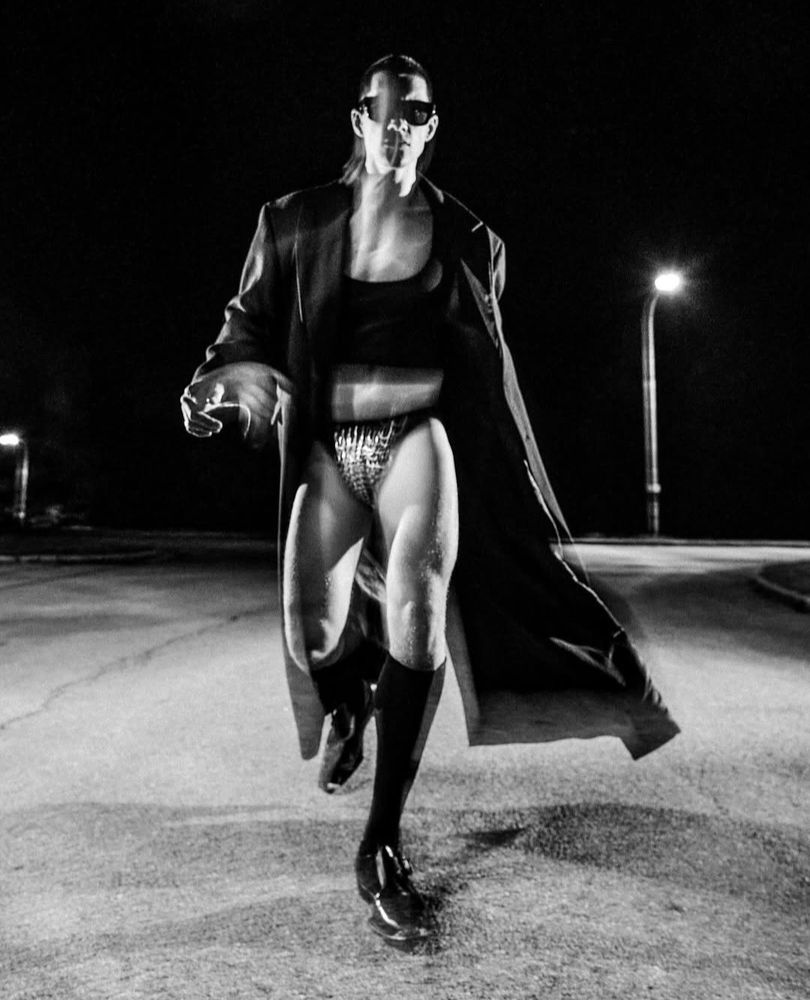Black and white, Model Augusta walking down the street wearing a long coat, cropped black tank top, black briefs with metal details and shows with long socks