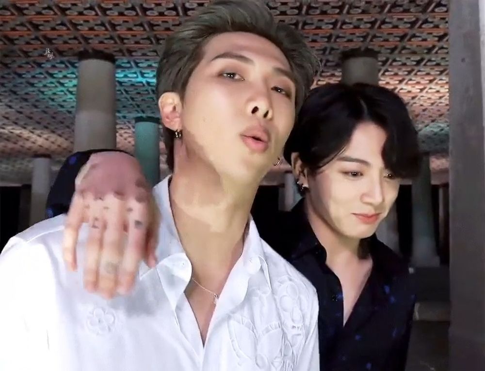 BTS's Namjoon and Jungkook during the filming of the Tonight Show Mikrokosmos performance. Namjoon has dark blind hair and a white dress shirt with white on white patterns on it. Jungkook has dark hair and a dark blue shirt. He has his arm sling around Namjoon, tattooed hand showing, and is smiling and looking down as if he has a secret. Namjoon is mid-expression, lips rounded in an "o.'