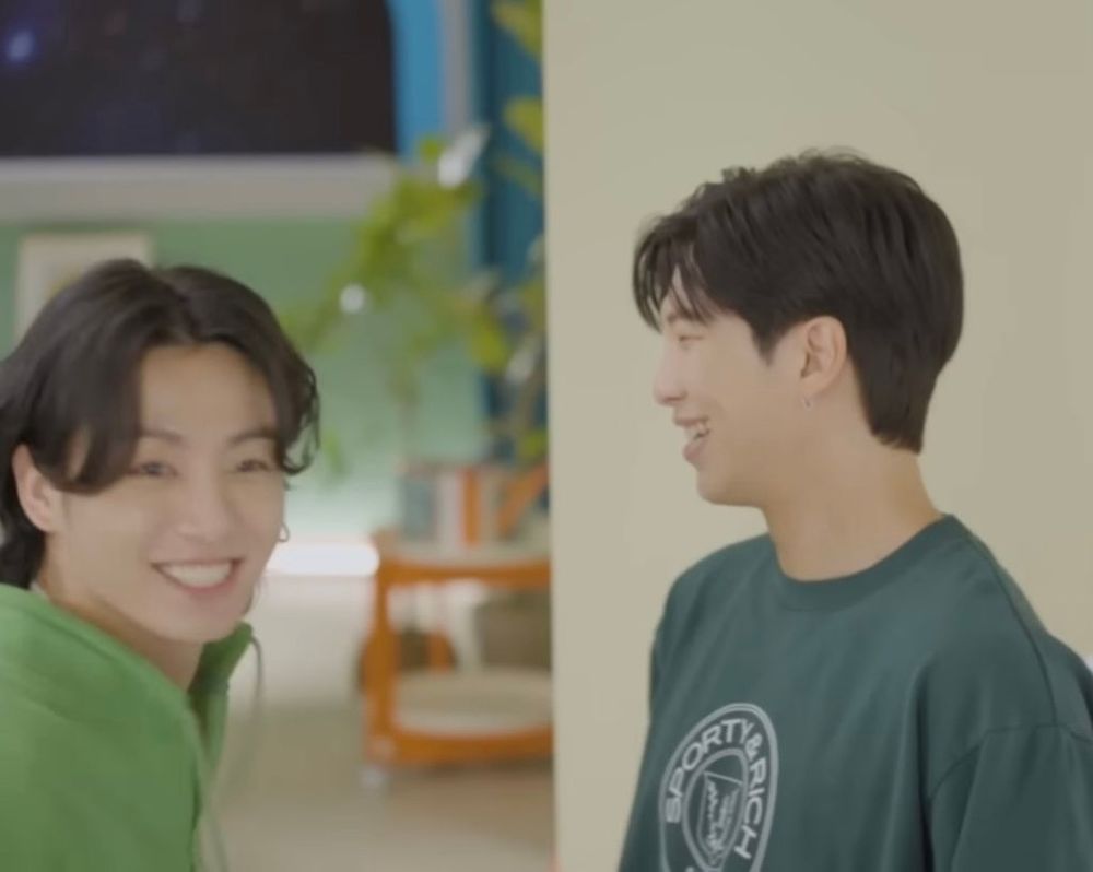 BTS's Jungkook and Namjoon, in a still from a video. Their standing opposite each other, but each is looking off to the right and smiling/laughing. Jungkook has a big big smile and his bangs are curling in a silly way. It's super adorable.