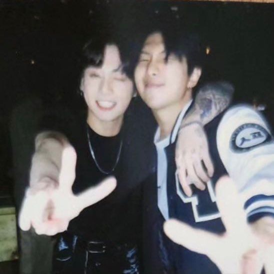 A blurry photobooth photo of BTS's Jungkook and Namjoon. Jungkook is in a black T-shirt and Namjoon is in a letterman's jacket. They both have floppy black bangs. They're winking and giving the camera peace signs. Jungkook has his tattooed arm slung around Namjoon's neck.