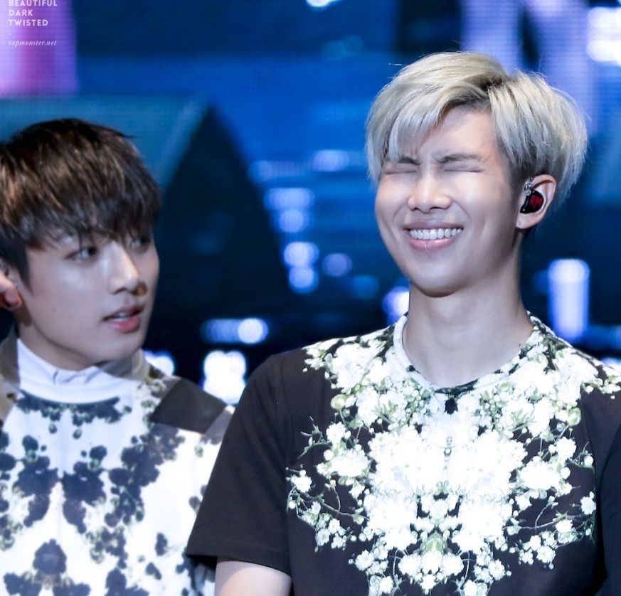 A young Jungkook with dark hair) and Namjoon (blond) in dark outfits with very shiny accents. Namjoon is smiling/laughing so hard his eyes are closed, and Jungkook is looking off to one side from beside and a little behind Namjoon.