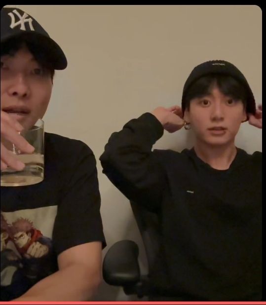 BTS's Namjoon and Jungkook sitting together on a weverse livestream. Namjoon is wearing a Yankees ball cap and a T-shirt and drinking something from a glass. Jungkook is wearing a long sleeved black shirt and is adjusting his black beanie. He has a lip piercing and an earring in.