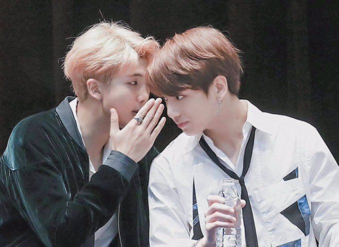 Namjoon, with blond hair, sitting next to Jungkook, who has brown hair. They're leaning into each other and Namjoon is whispering into Jungkook's ear behind one hand while Jungkook listens attentively.