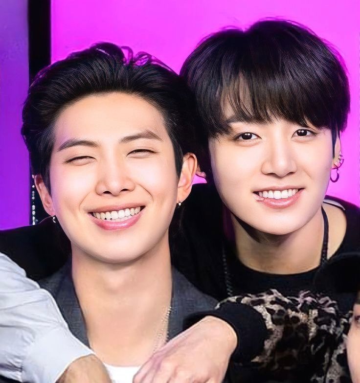 A closeup of BTS's Namjoon and Jungkook, heads close together, in front of a pink and purple background. Both have black hair and are smiling at the camera, Namjoon so hard his eyes are nearly closed. 