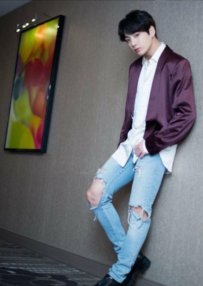 BTS's Jungkook leaning against a wall next to a brightly colored painting. He's wearing distressed jeans with the knees all but torn out and an untucked white dress shirt under a silky burgundy jacket. He's standing with one knee bent and is looking inquiringly into the camera.