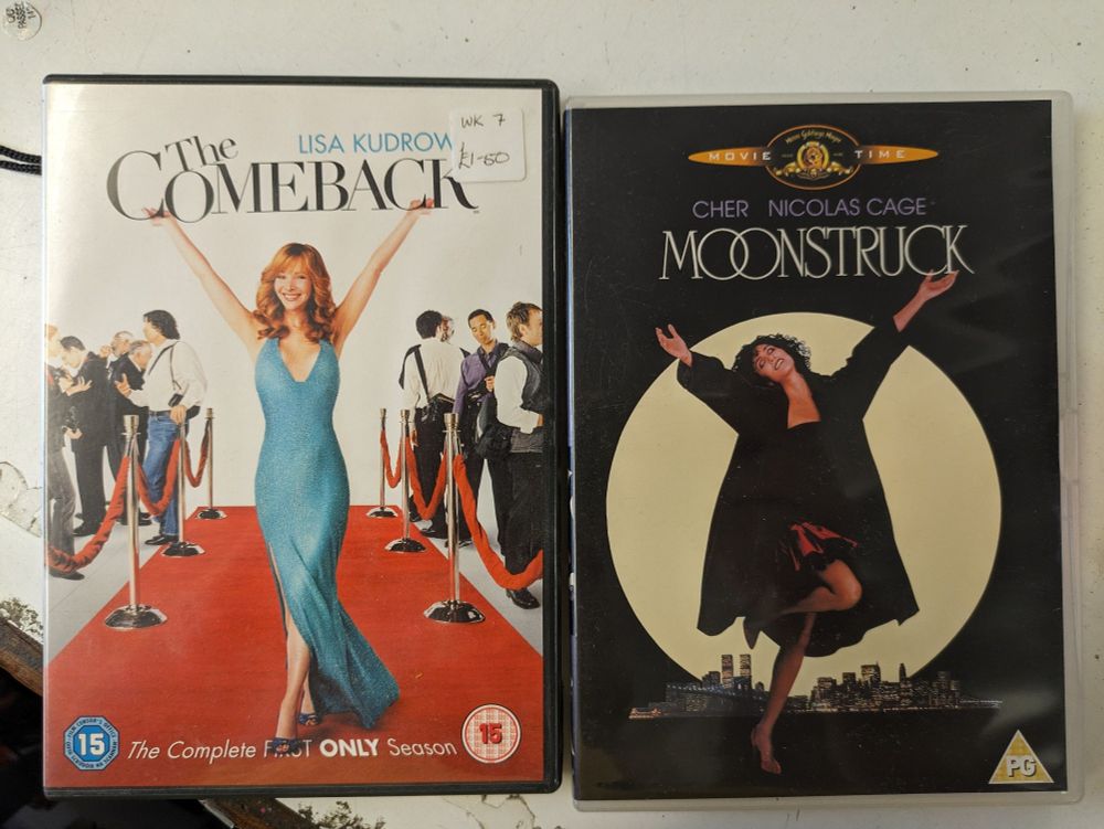 DVDs of The Comeback and Moonstruck, both covers featuring the lead character in an arms up, "here I am, life is amazing" pose