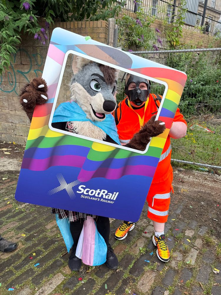 a wolf fursuiter wearing a trans pride flag and a person in a pup hood and high-visibility orange clothing pose together holding a rainbow-colored scotrail photo frame at glasgow pride 2025, standing on a cobbled path with confetti scattered on the ground.