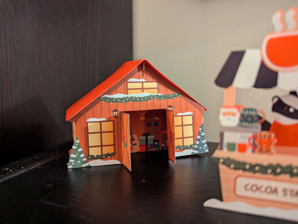 A colorful pop-up house with winter holiday decor. There are wrapped gifts inside. Benny is also in the foreground, waiting for customers to visit his cocoa stand.