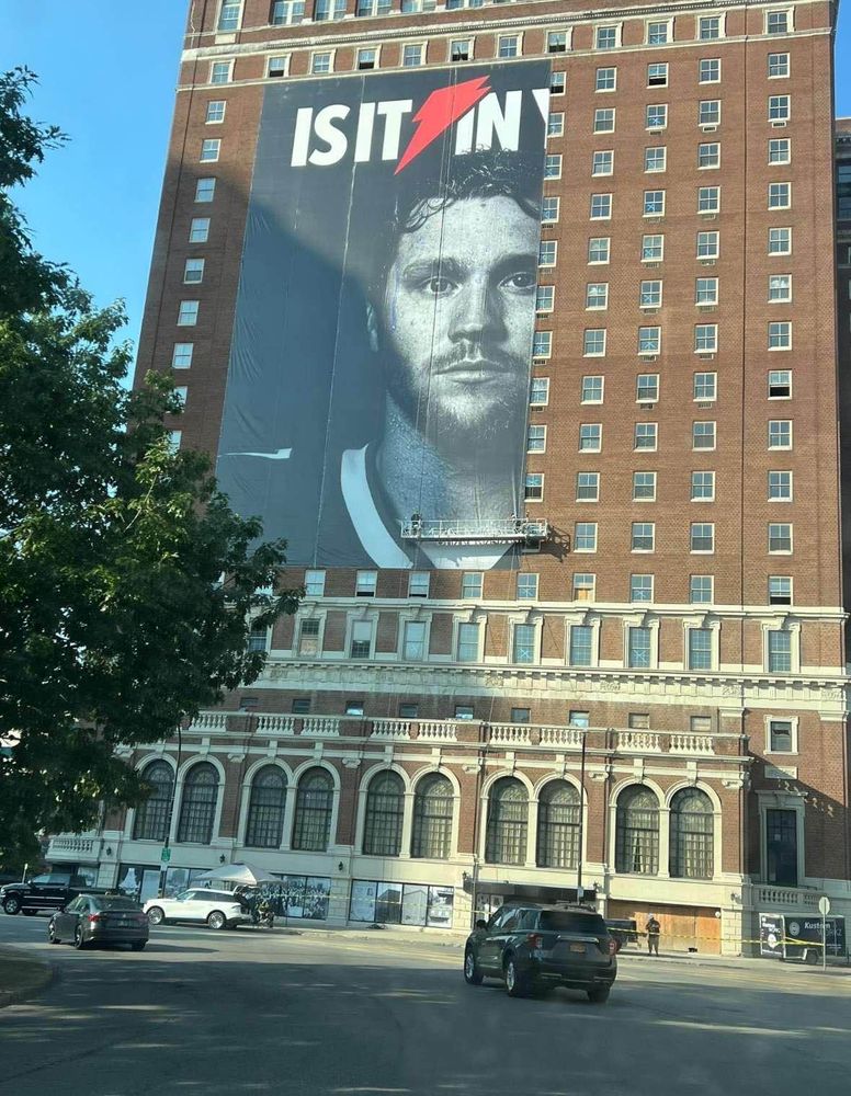 An advertisement in the process of being placed on the side of a building. It's a Gatorade advertisement with known NFL "First quarterback ever" JOSH FUCKING ALLEN of the Buffalo Bills stoically looking out to the streets in black and white. However, only 3/4 of the advertisement has been placed so the text says "Is it in" instead of "Is it in you"