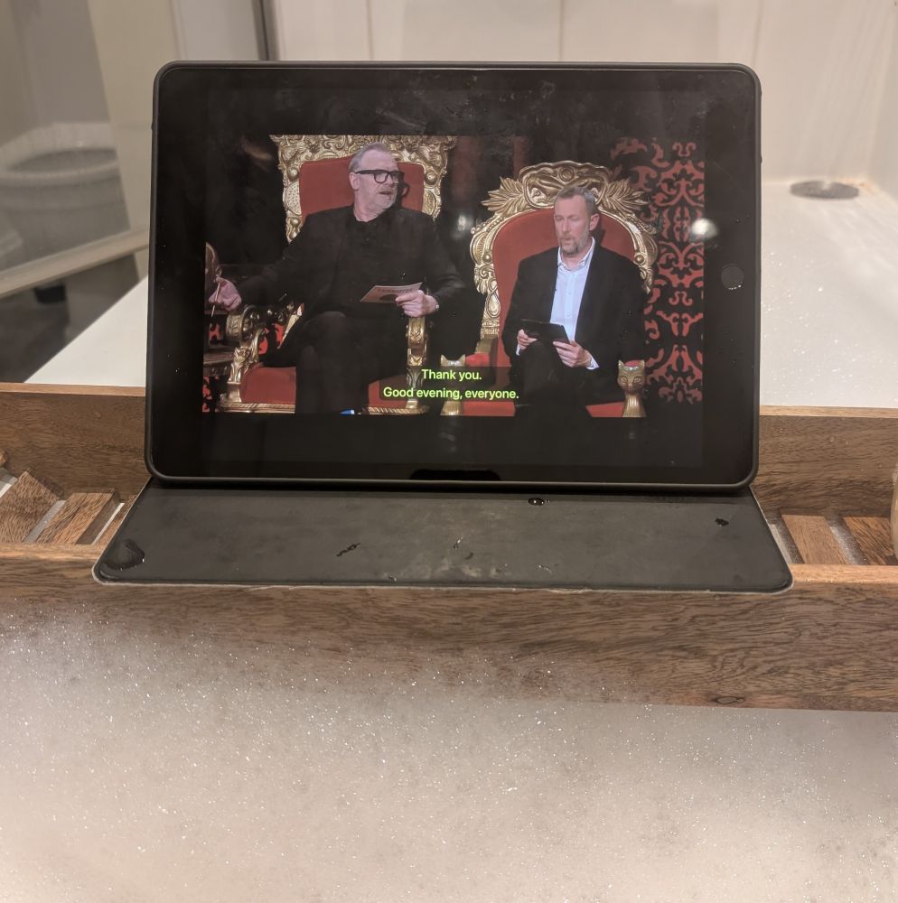 A wooden stand over a bath full of bubbles, with a tablet showing Greg Davies and Little Alex Horne on Taskmaster 