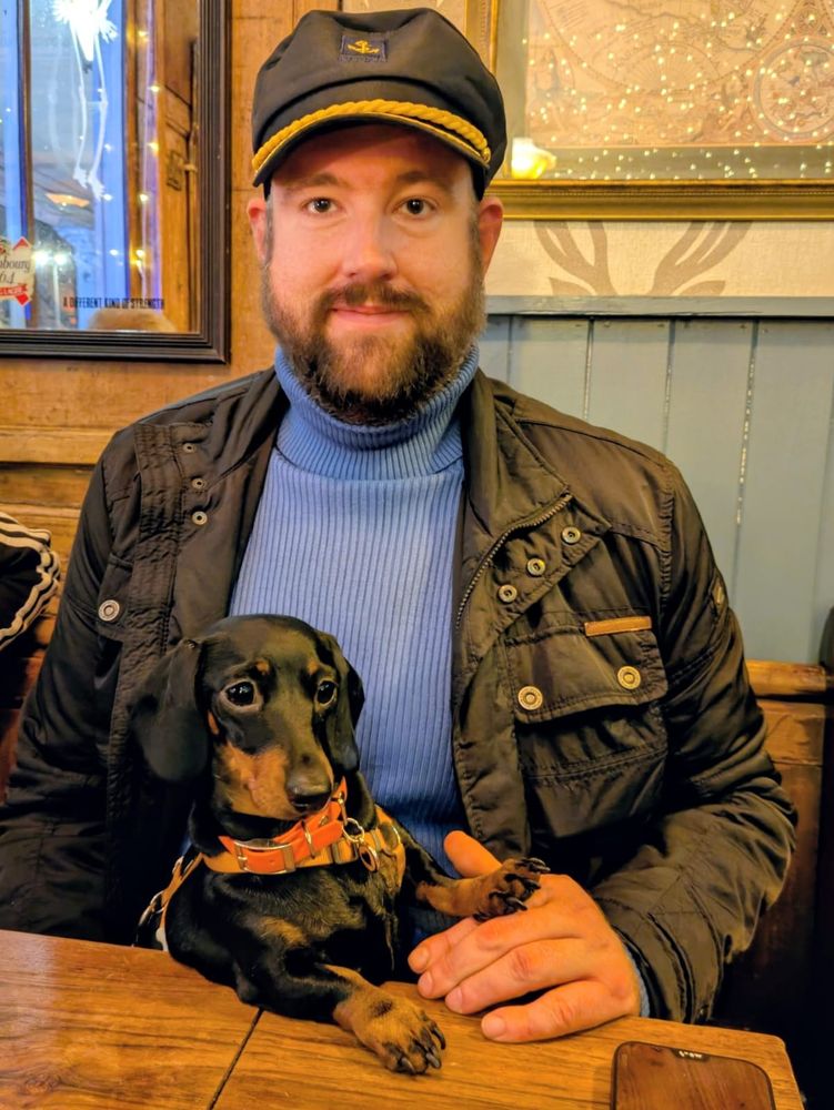 A man with dark coloured beard, in a blue turtleneck, sailors hat, and black work coat, holding a black and tan miniature dachshund in repose. 