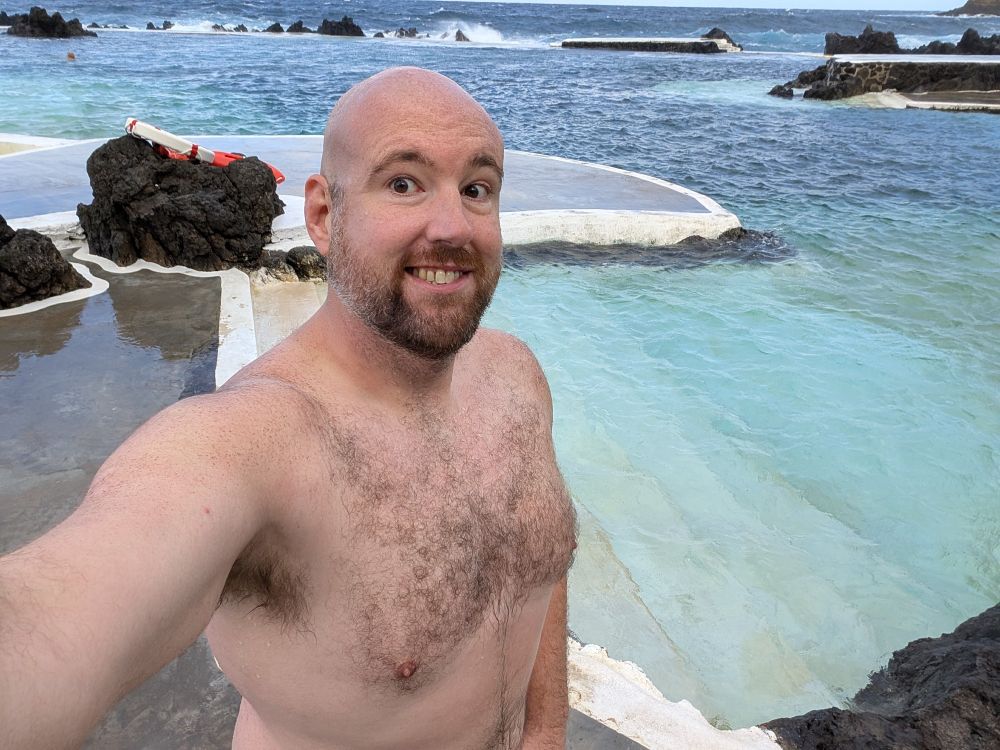 A bald white man with beard and hairy chest, grinning standing outside of an ocean pool. 