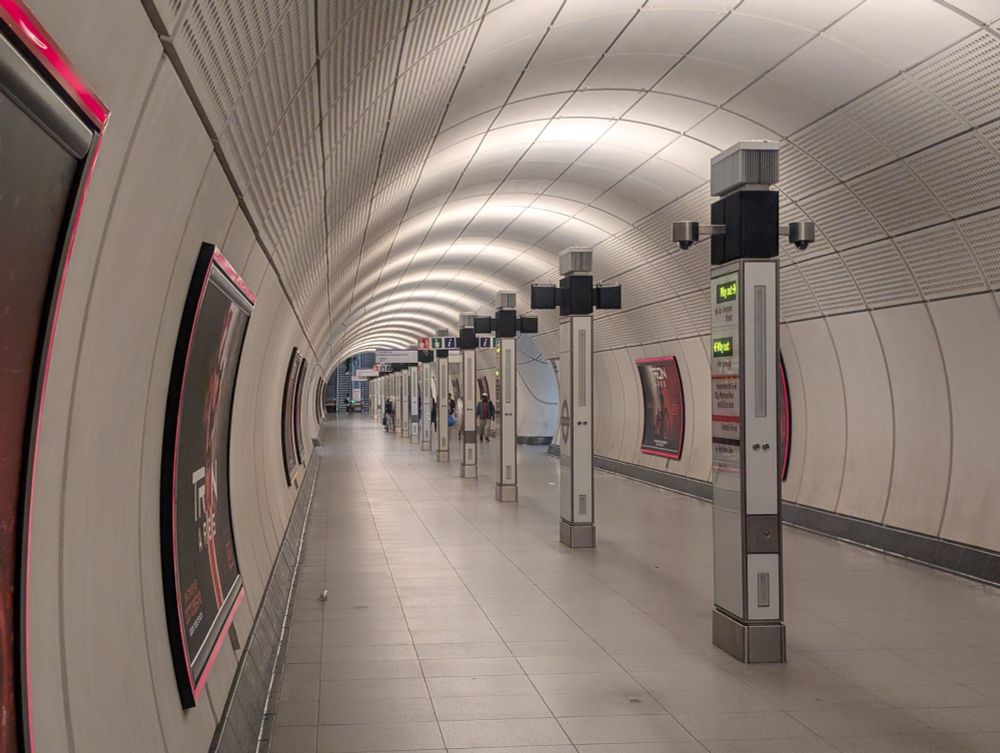A gray concrete Elizabeth Line station tunnel, mostly empty of passengers, with rings of white light and a series of white and silver plinths toppes with black fixtures. 