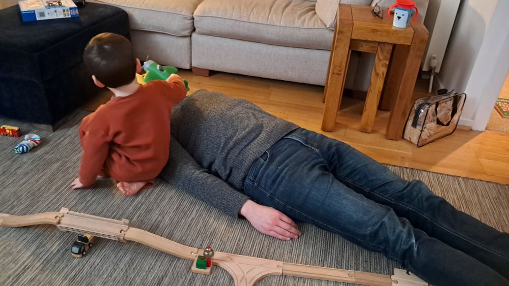 Toddler playing with a toy aeroplane on his father who is lying flat on his back on the floor
