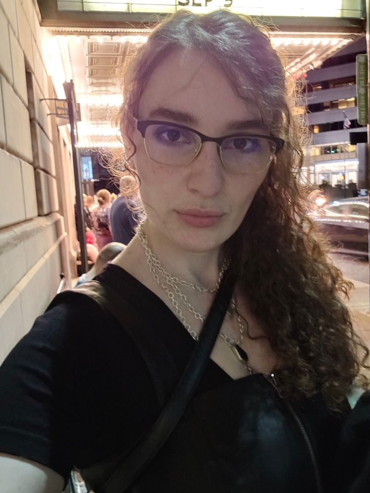 Em with glasses, outside without a mask and the theatre with Marquis lights shining behind. Her bag strap is crossing their chest. There are silver chains leading to a black coffin pendant.