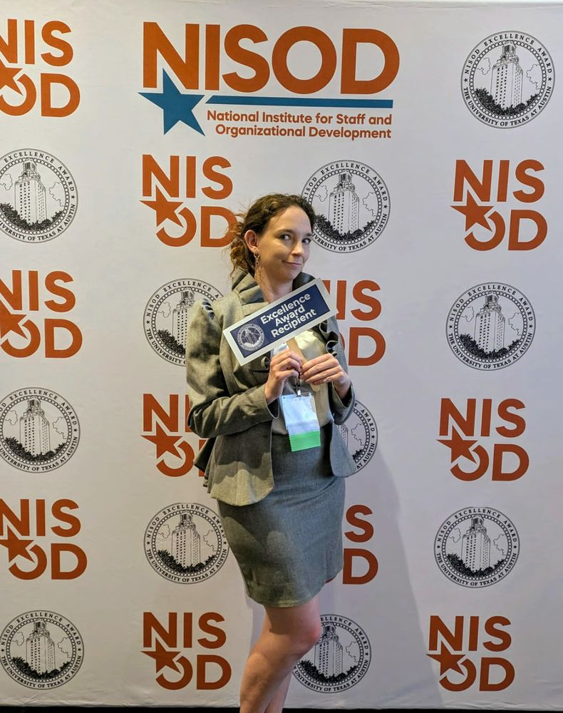 A woman in a gray ruffled suit standing in front of a white backdrop with NISOD in red font. She is holding a sign that says "Excellence Award Recipient."