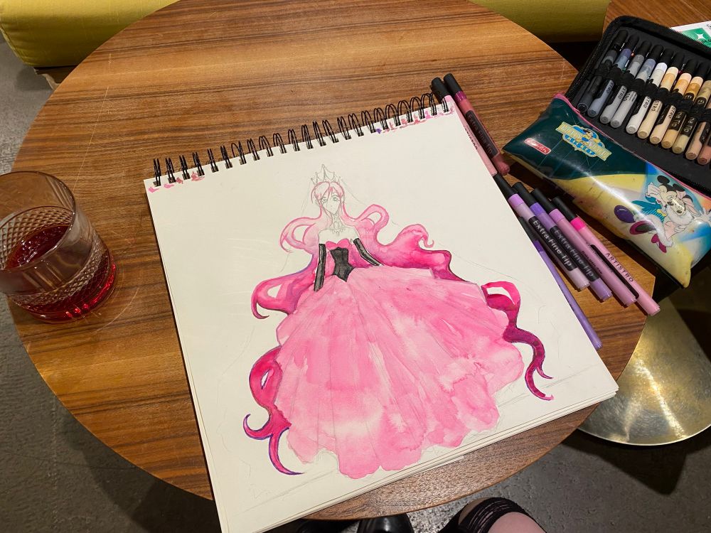 A huge drawing of a character that looks like a Princess! She is wearing a pink dress and has long pink hair! The drawing is halfway finished! The drawing is laying on a round wooden table and is surrounded by a drink in a glass and art supplies!