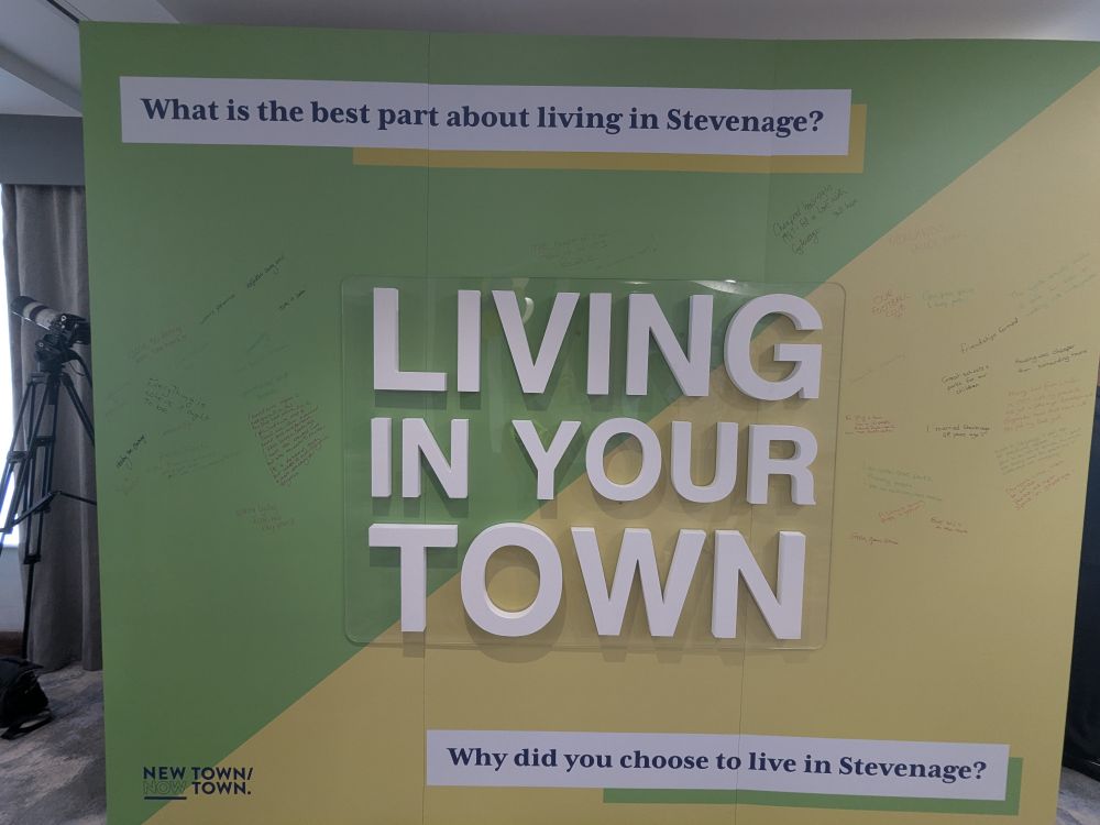 A green and yellow backdrop with white text that asks "What is the best part about living in Stevenage?" and "Why did you choose to live in Stevenage?" The text "LIVING IN YOUR TOWN" is prominently displayed in the center of the backdrop. The bottom left corner says "NEW TOWN/NOW TOWN". People have written messages on the board.