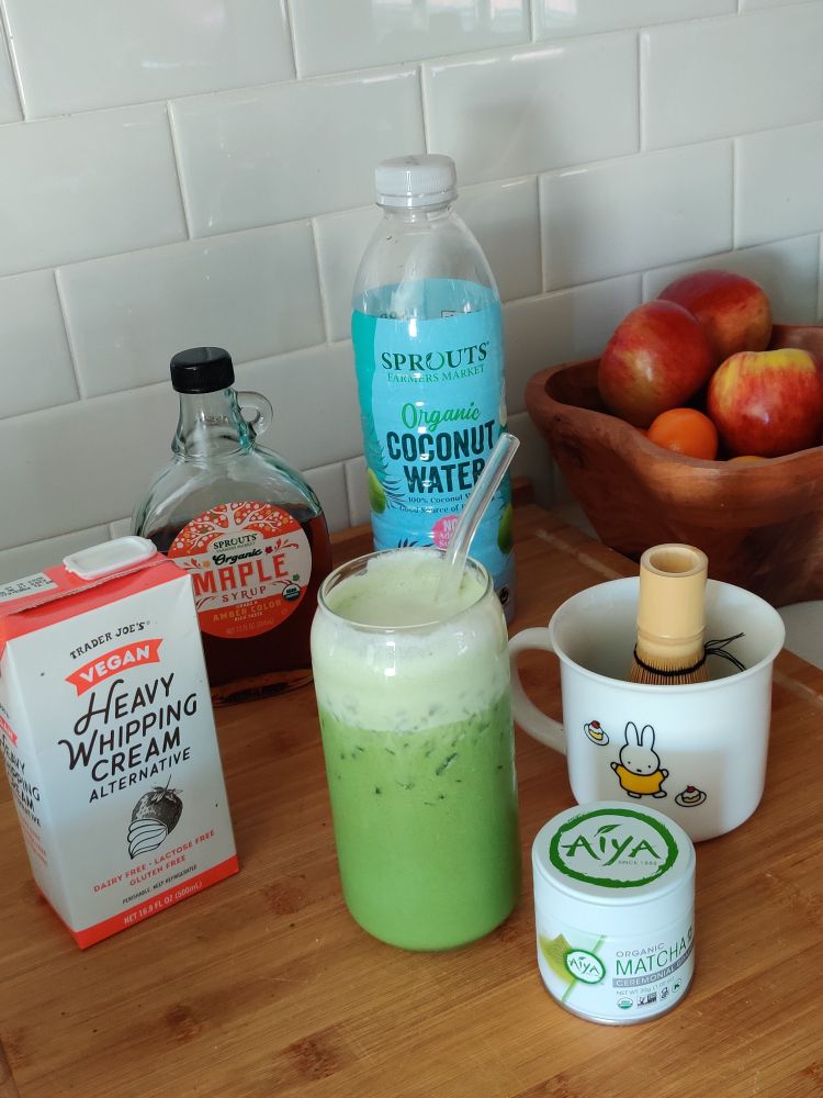 A photo of a cup of matcha and other ingredients next to it. Containers of trader Joe's heavy whipping cream, maple syrup, organic coconut water, and Aiya Matcha