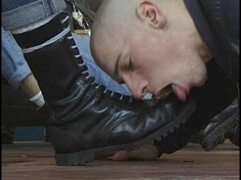 A person with a shaved head kneeling and licking a black leather boot