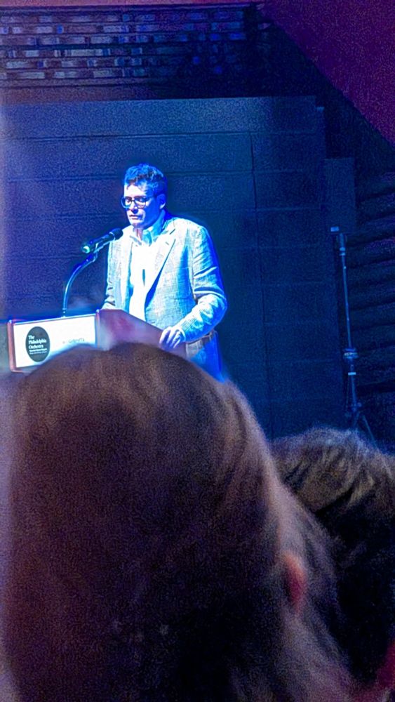 John Green as the end of the Philadelphia event for Everything is Tuberculosis, bathed in a blue light that makes him look ethereal, or like this was a photograph of a CRT Television. More audience heads are visible as well. 