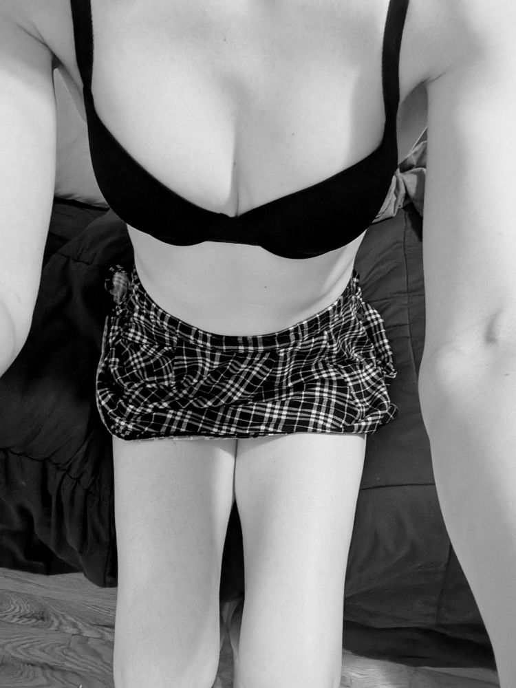 a black and white photo of a girl sitting on the edge of a bed on her bra and skirt