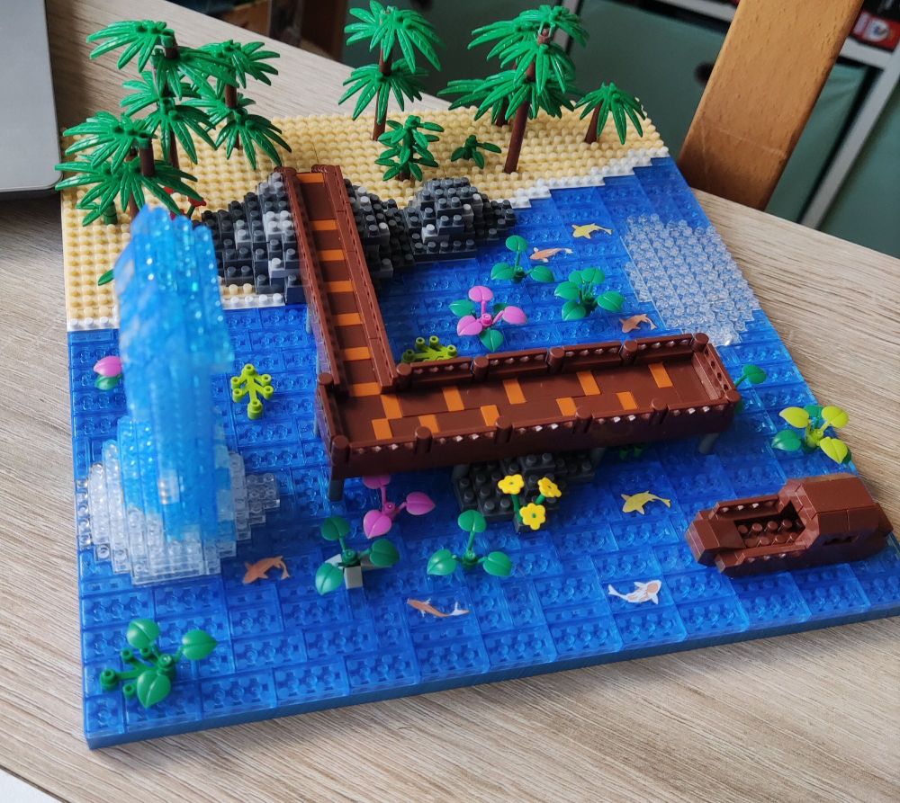 Szene aus Klemmbausteinen. Ein Steg ragt aufs Wasser hinaus, auf dem ein kleines Flusschiff fährt. Im Wasser schwimmen bunte Koikarpfen und Wasserpflanzen. Der Steg endet an einem winzigen Strand auf dem Bambus wächst. Aus dem Wasser ragt eine Säule nach oben, die mal einen Wasserfall darstellen wird.