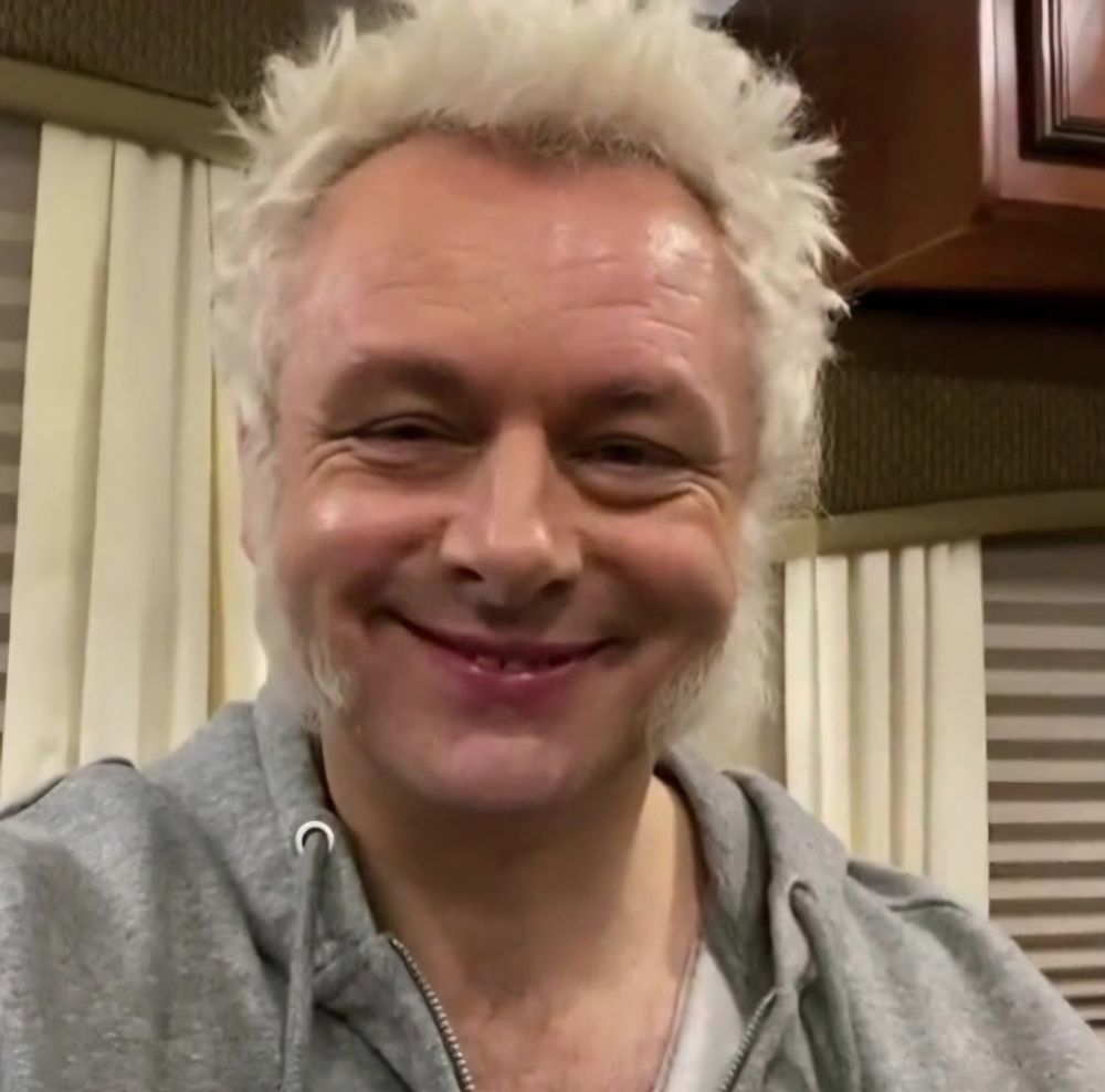 A smiling and beautiful Michael Sheen with Aziraphale's hair and mutton chops and wearing a grey hoodie.