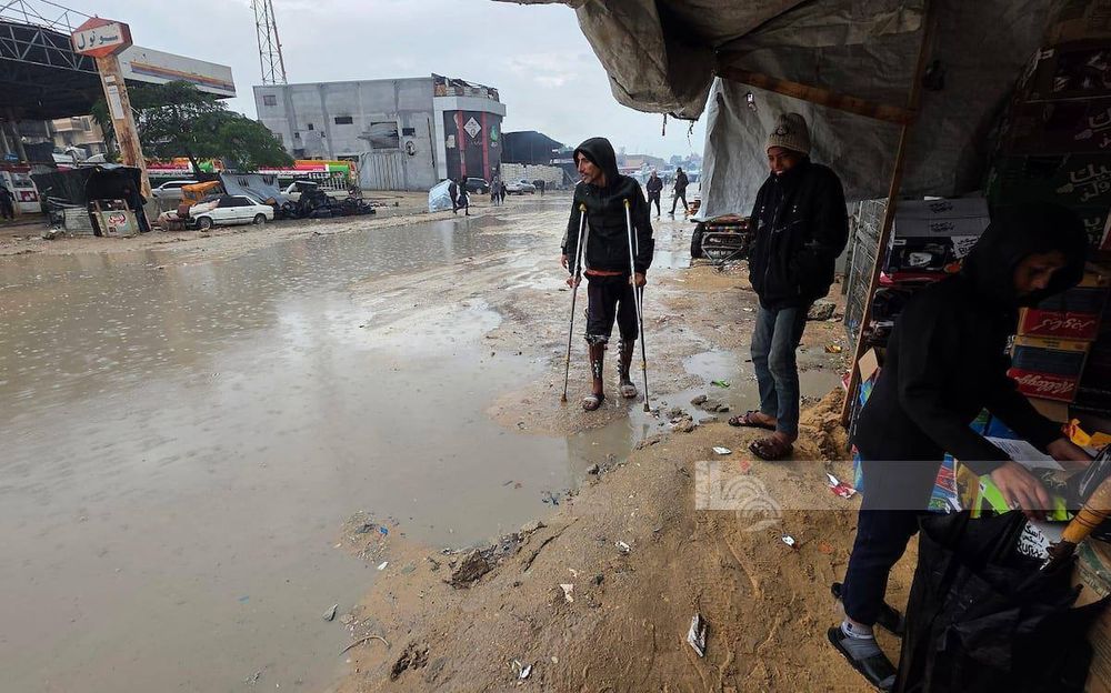 A man standing on crutches in a flooded, muddy street during heavy rain in Gaza. Several people stand nearby under a piece of tarp for shelter. Damaged cars and buildings appear in the background, showing the harsh winter conditions.