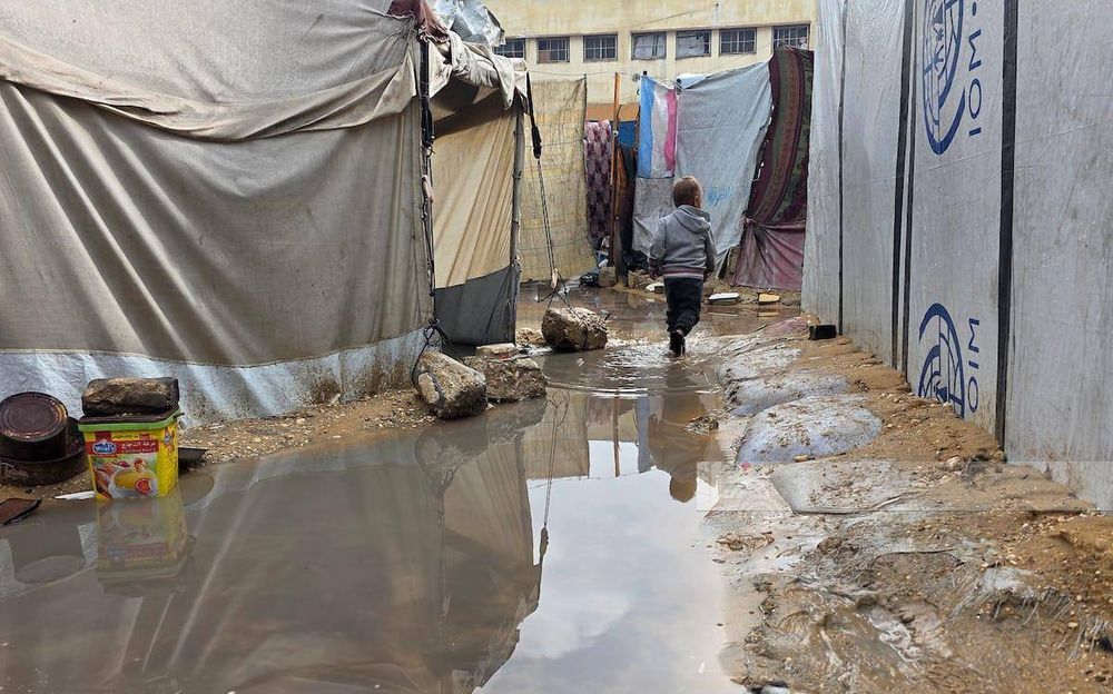 A small child walking through large puddles of water between worn-out tents tied with ropes and weighed down with rocks. The muddy ground and standing water show the difficult winter conditions in the camp.
