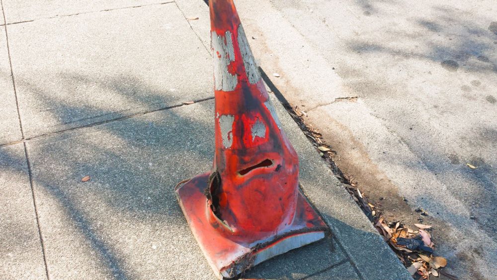 heavily used banged up scraped up traffic cone that looks like it's got 2 white eyes above a melted opening that is shaped like a surly mouth. It's giving Traffic Cone Foreman energy. 