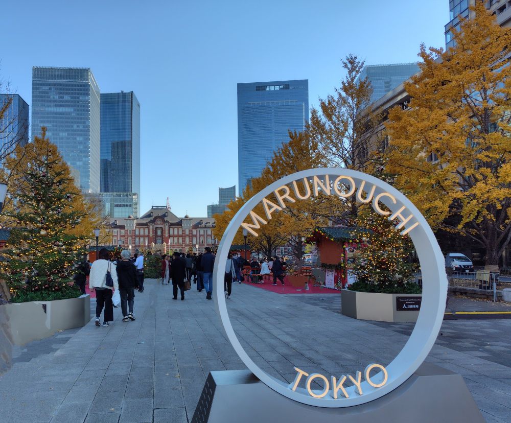 奥にビル群と煉瓦造りの東京駅。そこから道が伸びていて、両側に黄色に色付いたイチョウの並木。手前には、Marunouchi Tokyoという文字で飾り付けられた白い輪が置かれている。