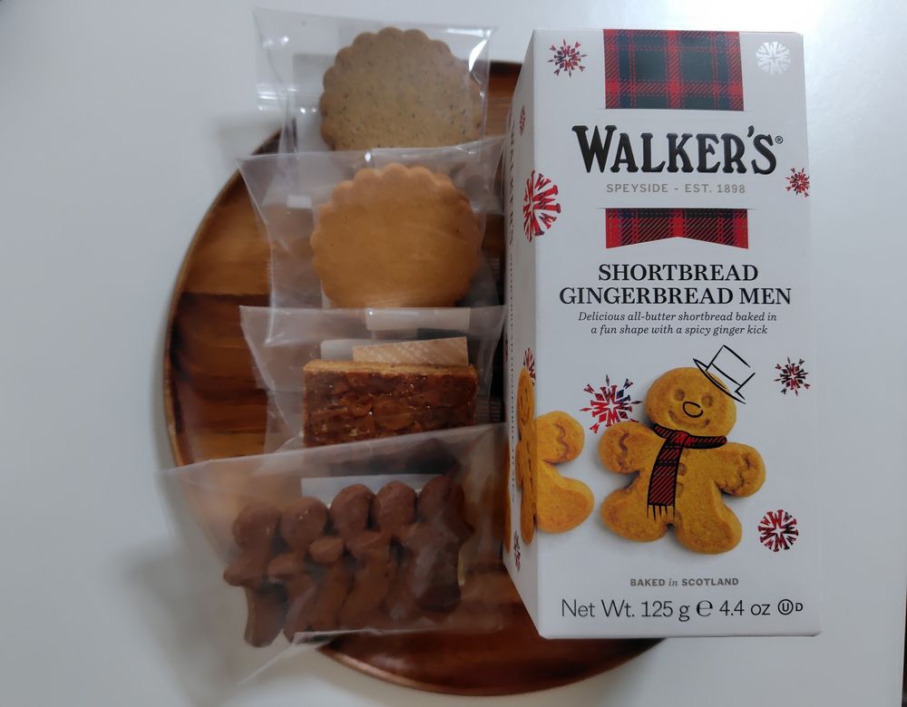 木の丸盆の上にクッキー数種。右側にウォーカーズのジンジャーブレッド・マンのクッキーが箱ごと。左側は4種のクッキーがそれぞれ小袋に入っている。上から花びら型のハニー・クッキー2種、フロランタン、小さなジンジャーブレッド・マンのクッキーが5枚。