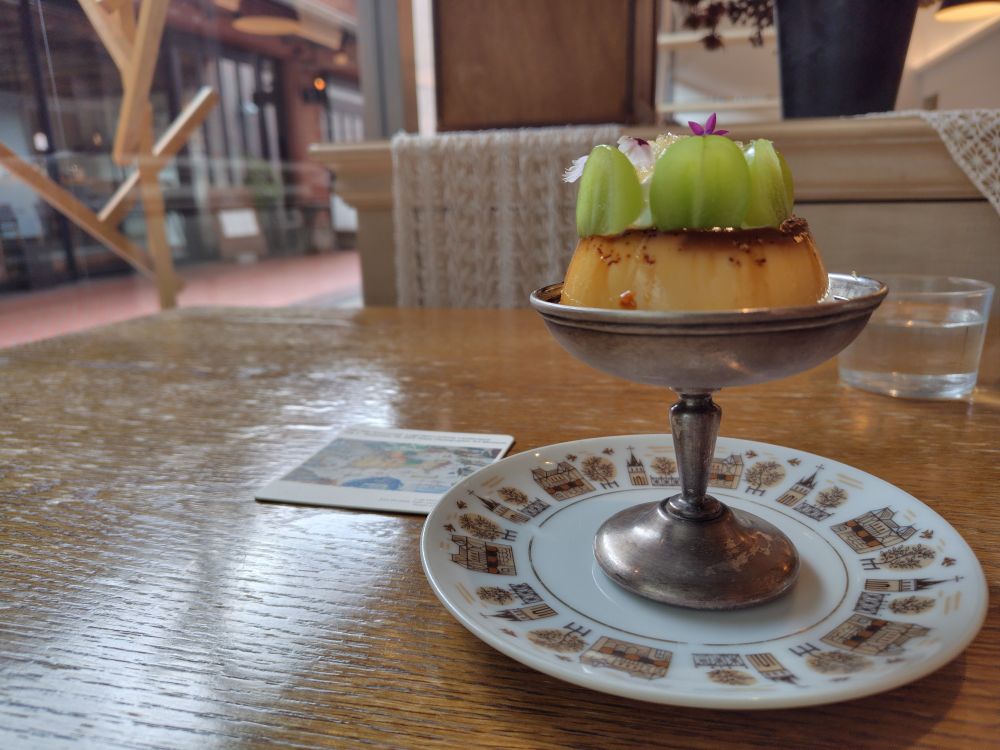 木のテーブルの上、やや右寄りに銀色の器に入ったプリン。その上には生クリームが載り、クリームを囲むように、カットされたシャインマスカットが並ぶ。