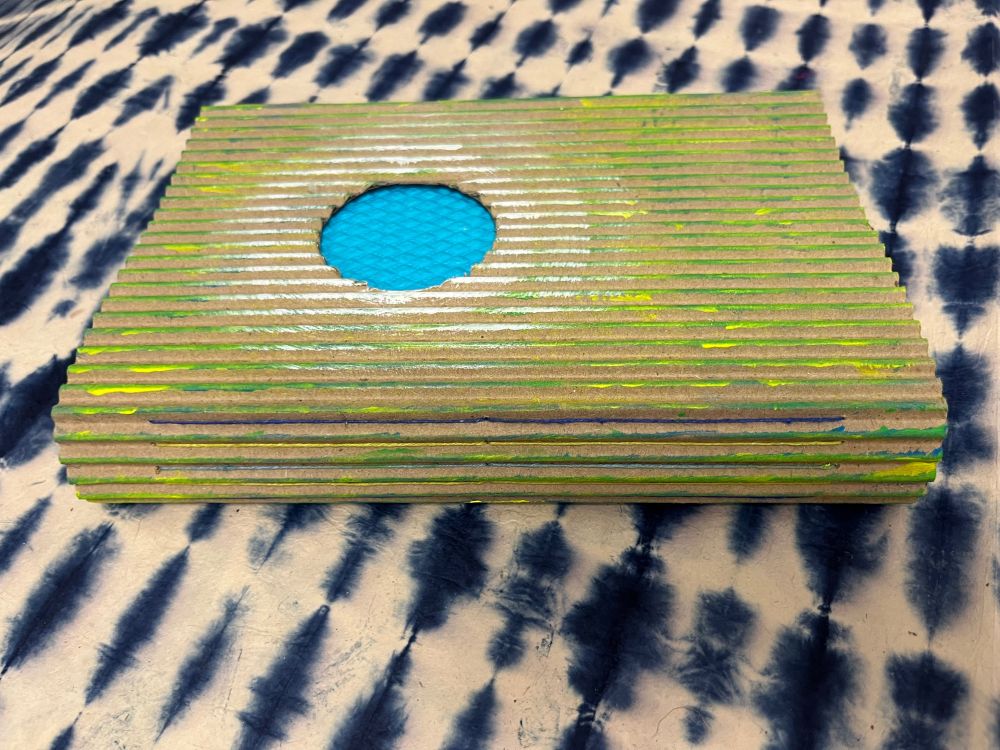 Handmade book. The cover is made from corrugated cardboard and is brushed in green, yellow, blue, and white paint. A circle is cut from the front cover, exposing the textured, blue paper underneath.